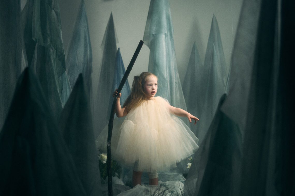 Kind in weissem Kleid in einer Kulisse von aufgehängten Tüchern.